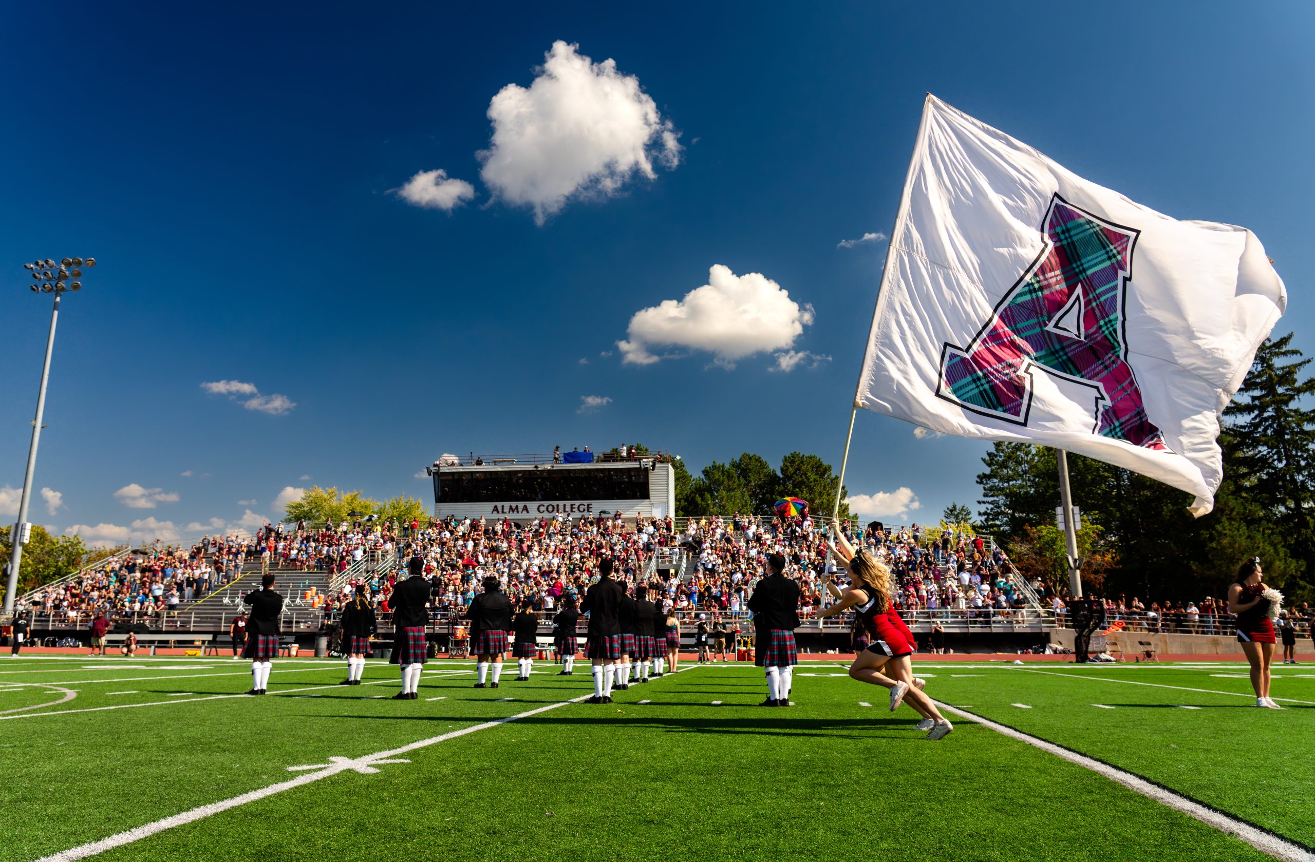 A cheerleader runs across a football field waving a large flag with a letter A as a marching band performs and alumni, along with other spectators, fill the stadium bleachers under a blue sky.