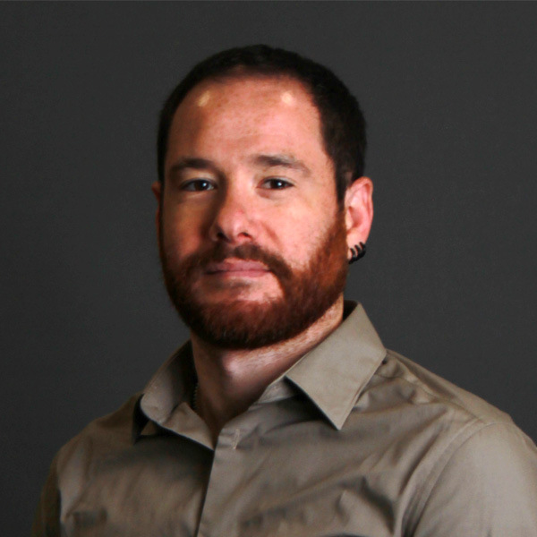 A man with short dark hair, a reddish beard, and earrings, wearing a tan collared shirt, stands against a dark gray background.