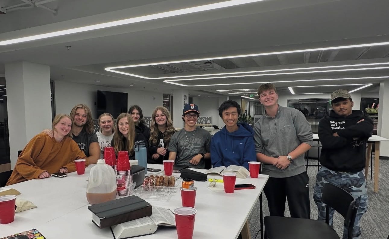 A group of ten young adults smile for a photo around a table with red cups, snacks, and books in a modern, well-lit indoor space.