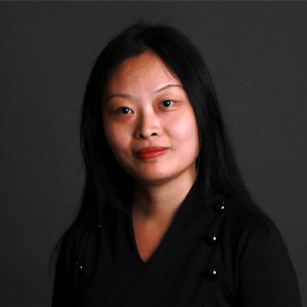 A woman with long black hair, wearing a black top, is posing in front of a plain dark background.