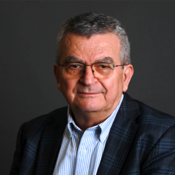 An older man with short gray hair and glasses, wearing a striped button-up shirt and a dark plaid blazer, sits against a plain dark background, looking at the camera—this is Recep Pekdemir.
