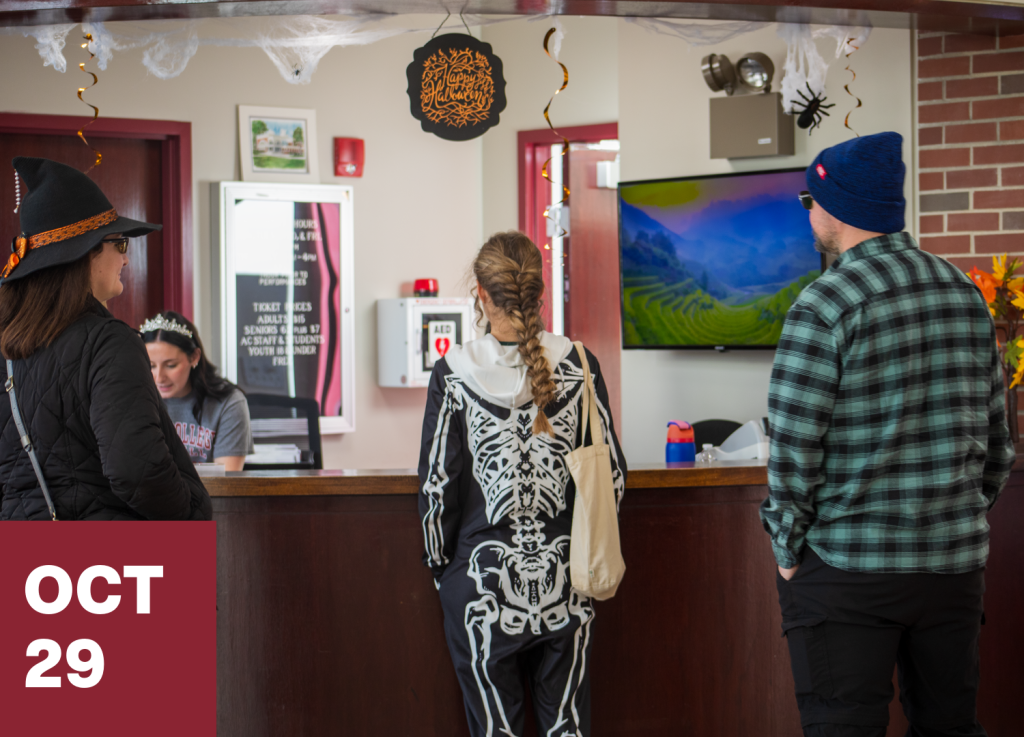 Three people in costumes stand at a reception desk decorated for Halloween, creating a festive setting perfect to connect-and-visit with others. A sign reads OCT 29 in the lower left corner. A woman behind the desk appears to be helping them. A TV and autumn leaves are visible.
