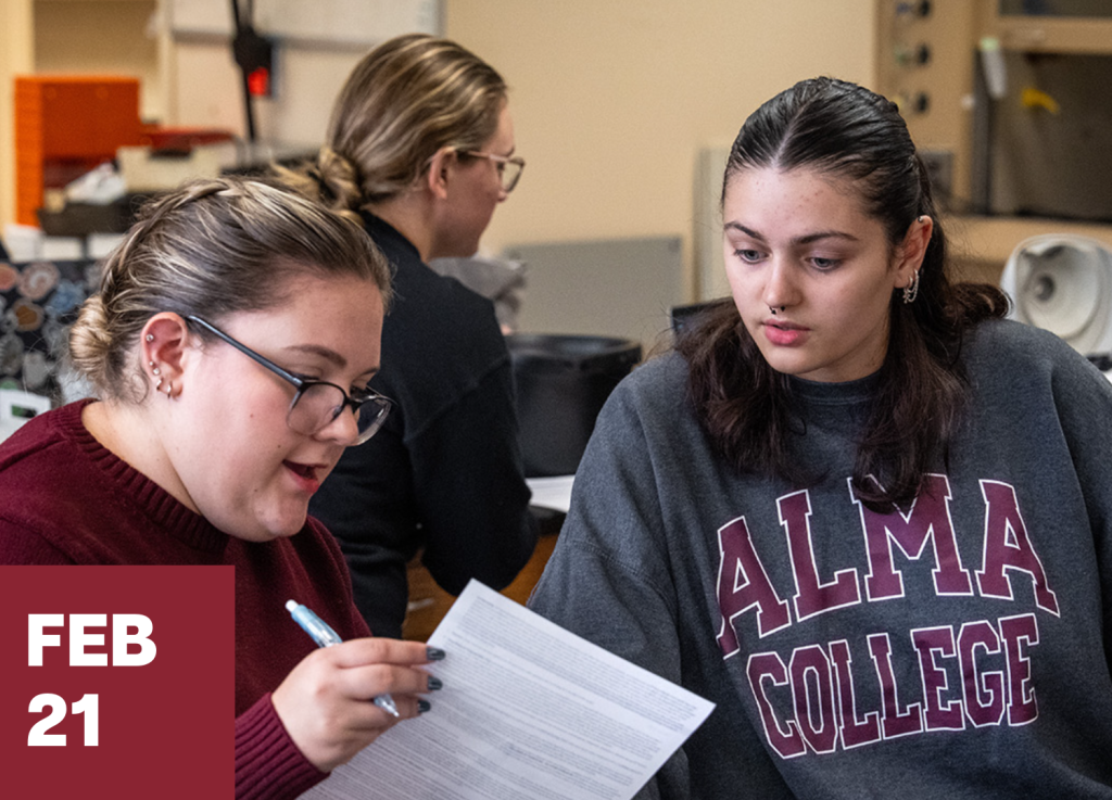 Two students in a classroom work together, one holding a paper and pen, while another listens. Both appear focused as they engage in a connect-and-visit activity. A date box in the corner reads Feb 21. Other students and equipment are visible in the background.