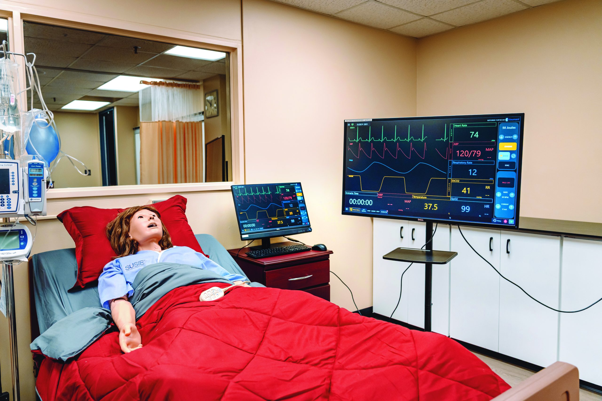 Medical simulation mannequin wearing a tartan hospital gown in a bed with vital signs displayed on two monitors.