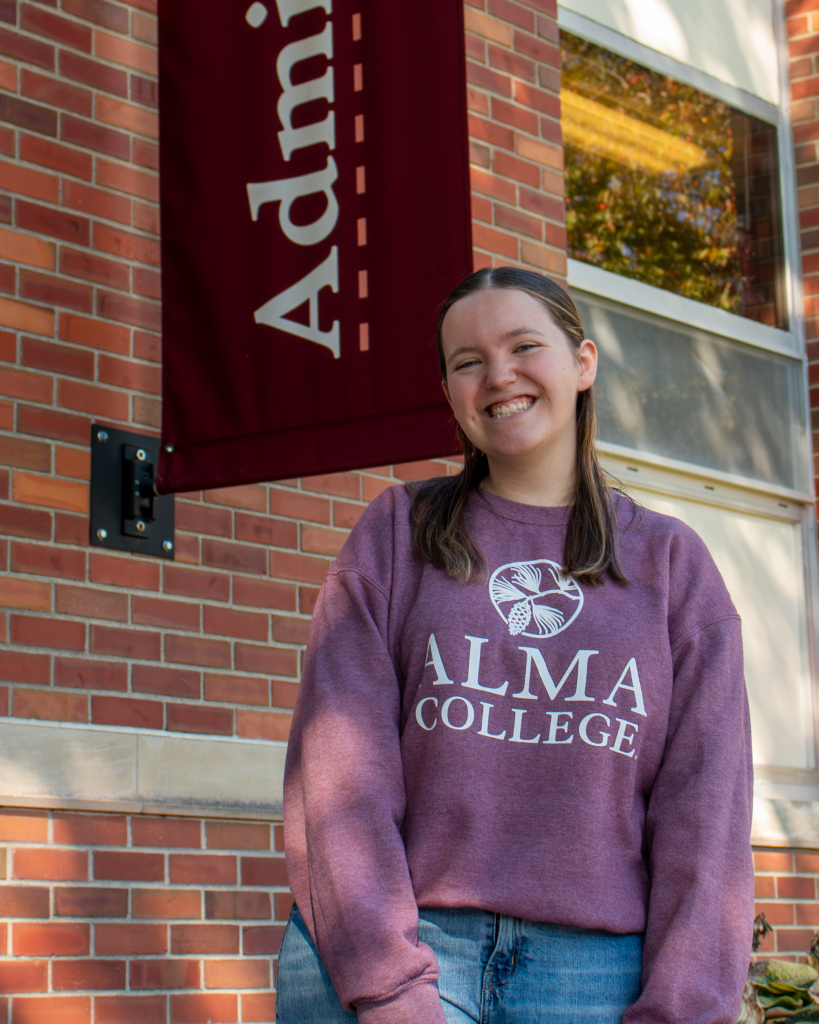 A smiling tour guide wearing a maroon Alma College sweatshirt stands in front of a brick building with an admissions banner.