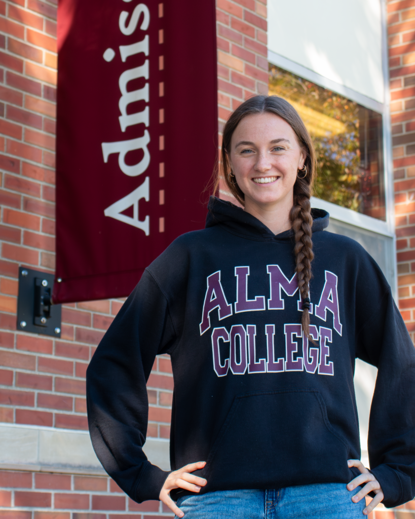 A tour guide wearing an Alma College hoodie stands in front of a brick building with a vertical maroon banner that reads Admissions.