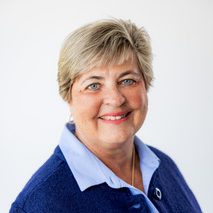 Smiling older woman with short gray hair, wearing a blue blazer over a light blue collared shirt, stands confidently in front of a plain white background—an inspiring presence among trustees.
