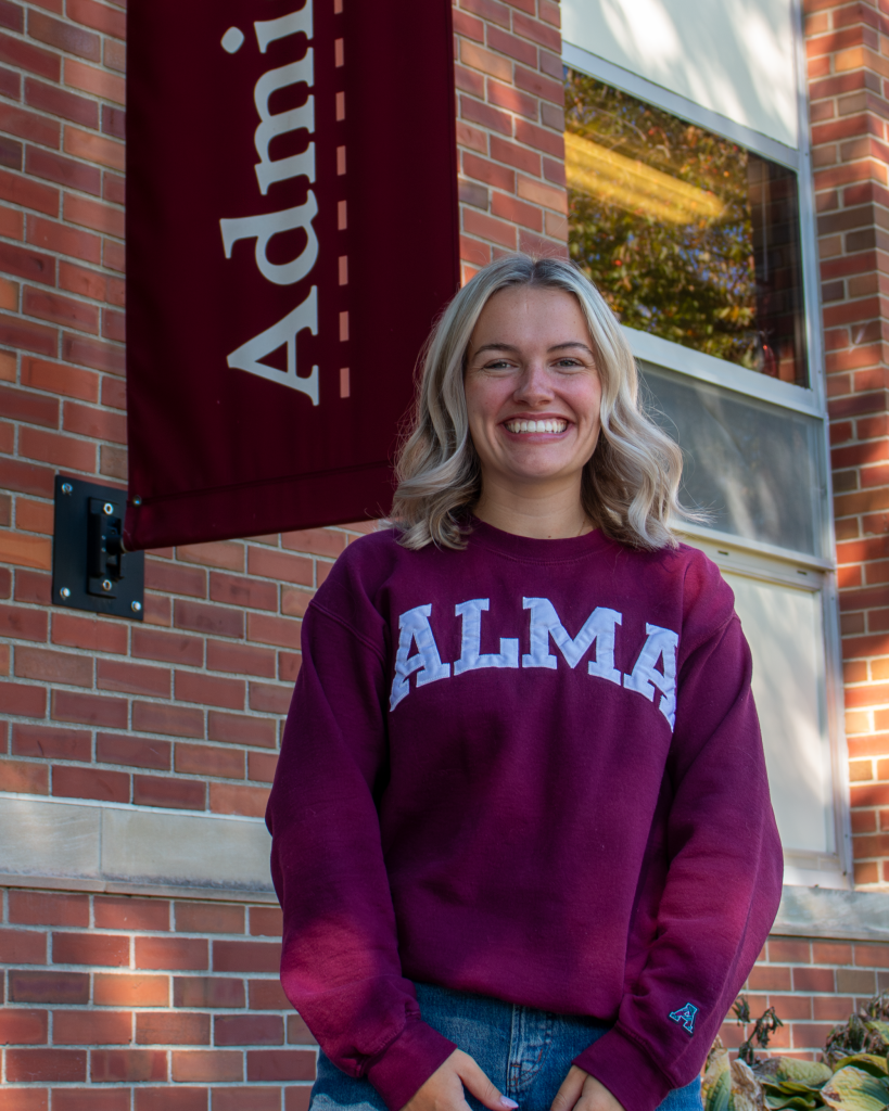 A smiling student tour guide wearing a maroon Alma College sweatshirt stands in front of a brick building with a maroon sign that reads Admissions