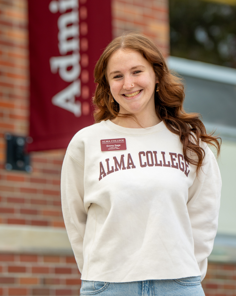 Emma Sage, an admissions ambassador and tour guide at Alma College, smiling while wearing a branded cream sweatshirt and a name tag.