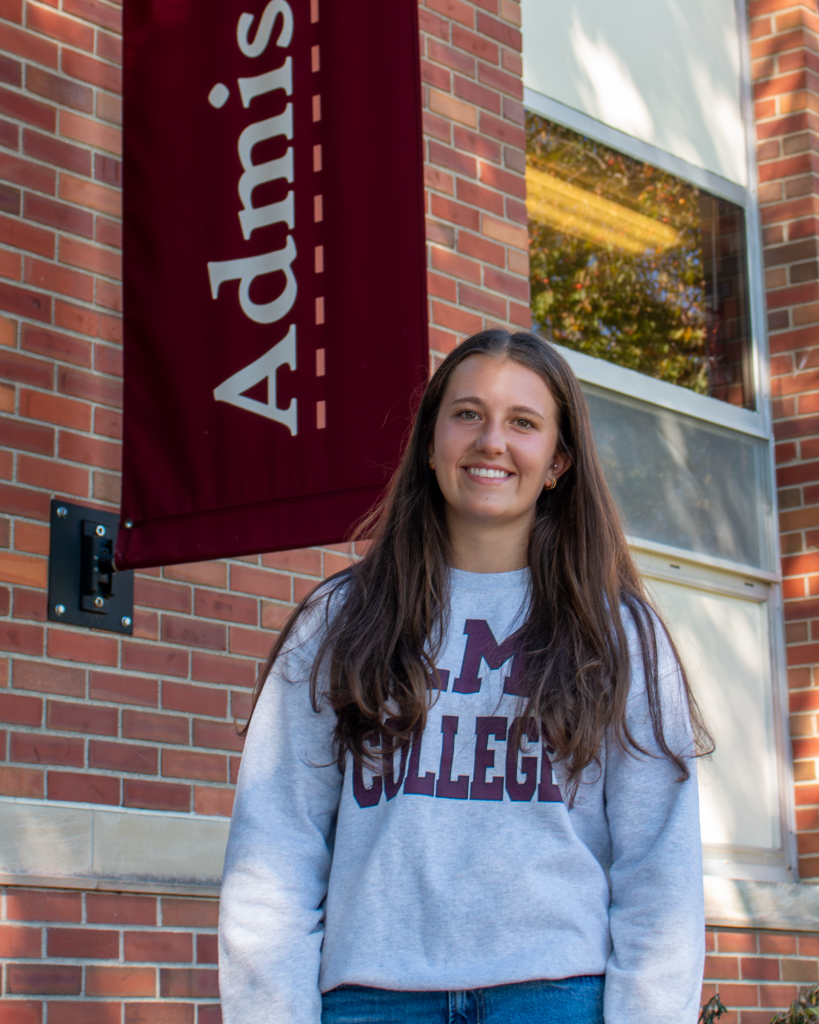 A smiling student tour guide wearing a gray college sweatshirt stands in front of a brick building with an Admissions banner.