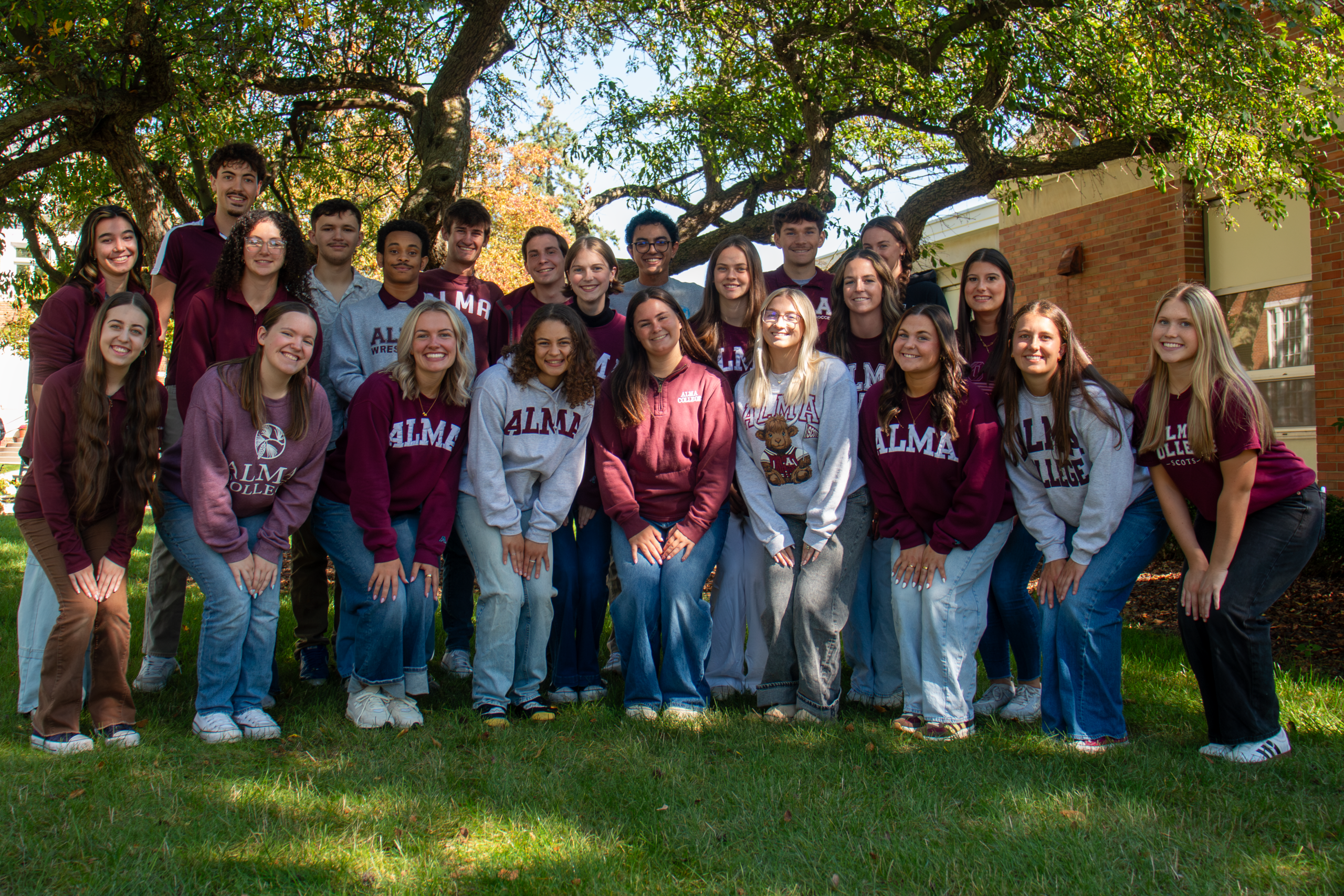 A large group of Alma College tour guides in branded sweatshirts and apparel posing for a photo outdoors under trees.