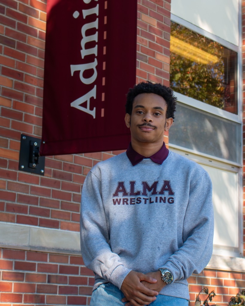 A young tour guide wearing a grey Alma Wrestling sweatshirt stands in front of a brick building with an Admissions sign.
