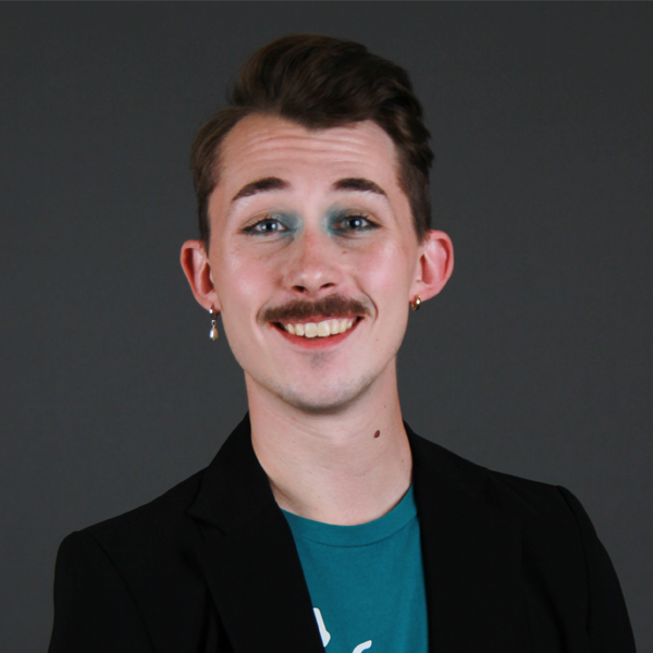 A person with a mustache and blue eyeshadow smiles at the camera, wearing a teal t-shirt under a black blazer as part of the Office of Belonging culture and leadership.
