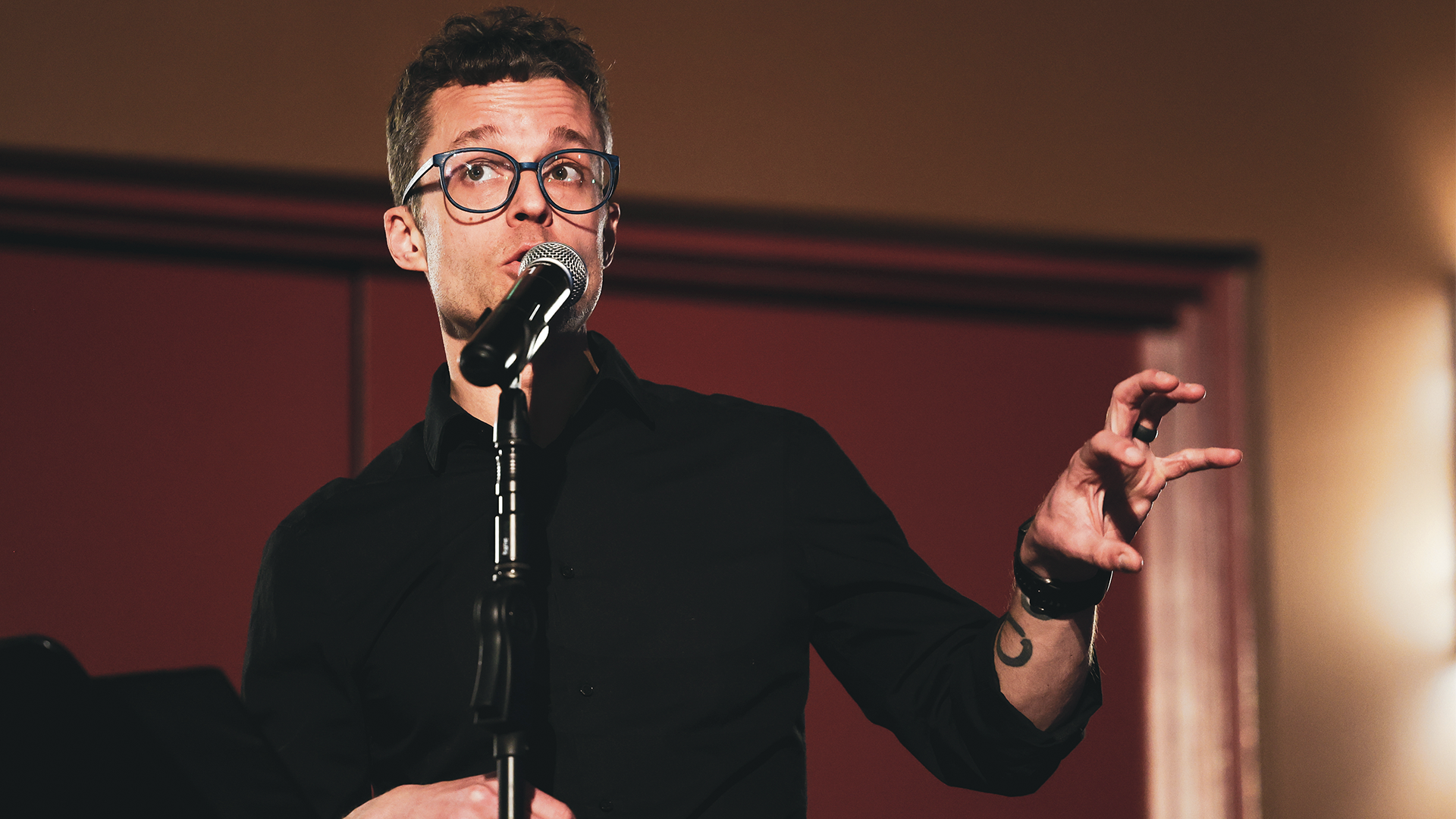 A composer with glasses speaks into a microphone while gesturing with his hand to choir students.