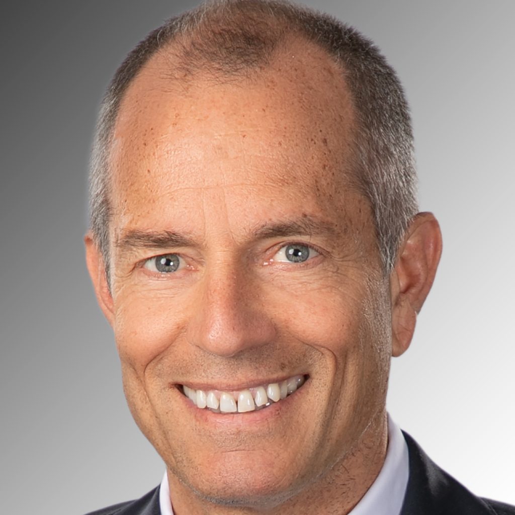 Smiling man with short graying hair, wearing a dark suit, white shirt, and light blue patterned tie, representing the trustees against a gray gradient background.