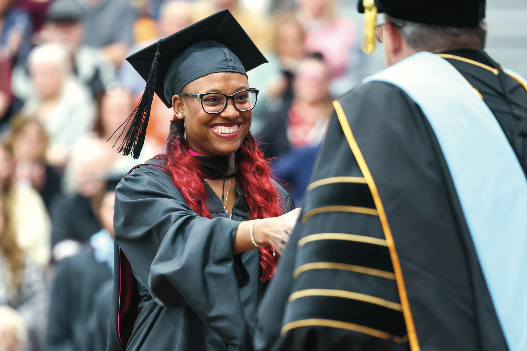 A smiling graduate in a cap and tartan gown shakes hands with a faculty member at a commencement ceremony.