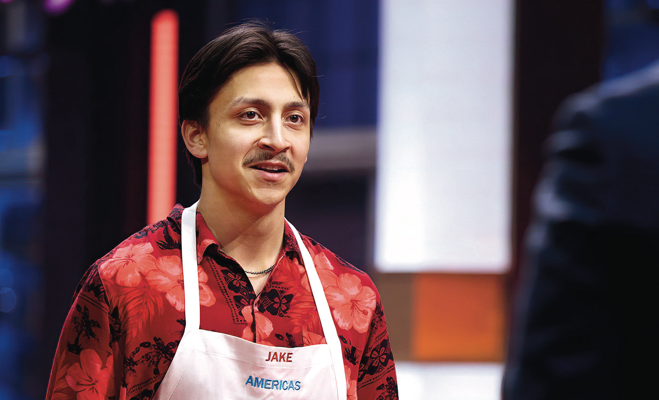 A MasterChef contestant named Jake wearing a red floral shirt and a white apron stands in a kitchen studio facing a judge.