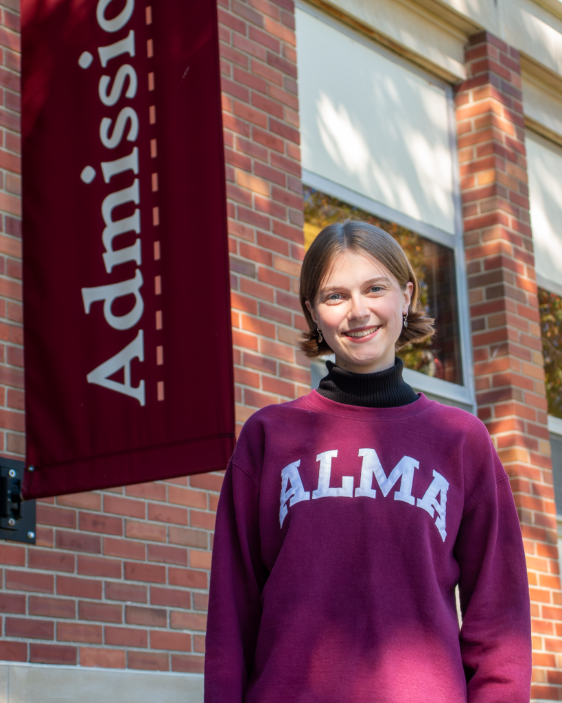 A student tour guide wearing a maroon Alma College sweatshirt smiles in front of a brick building with an Admissions banner.