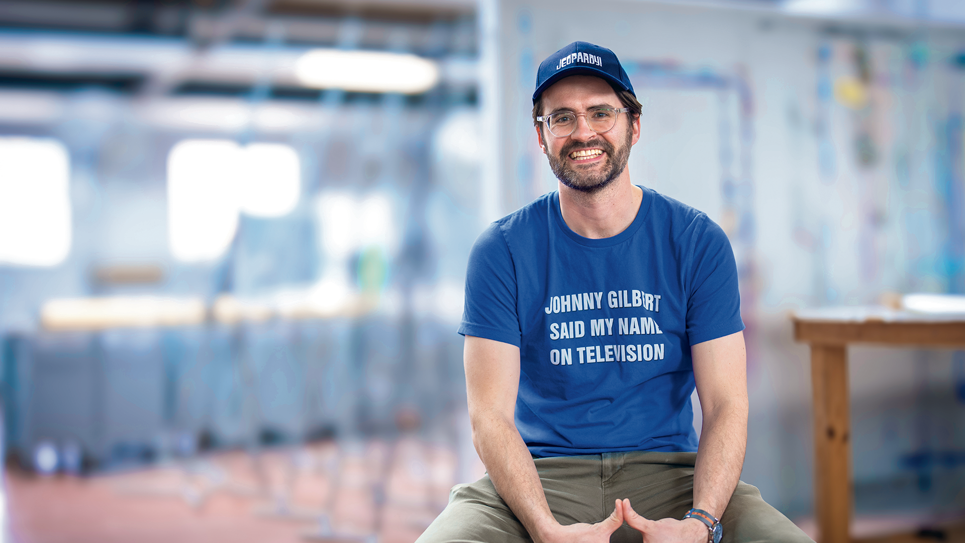 A smiling man wearing glasses, a Jeopardy! baseball cap, and a blue t-shirt that says Johnny Gilbert said my name on television as a second-chance-at-jeopardy.