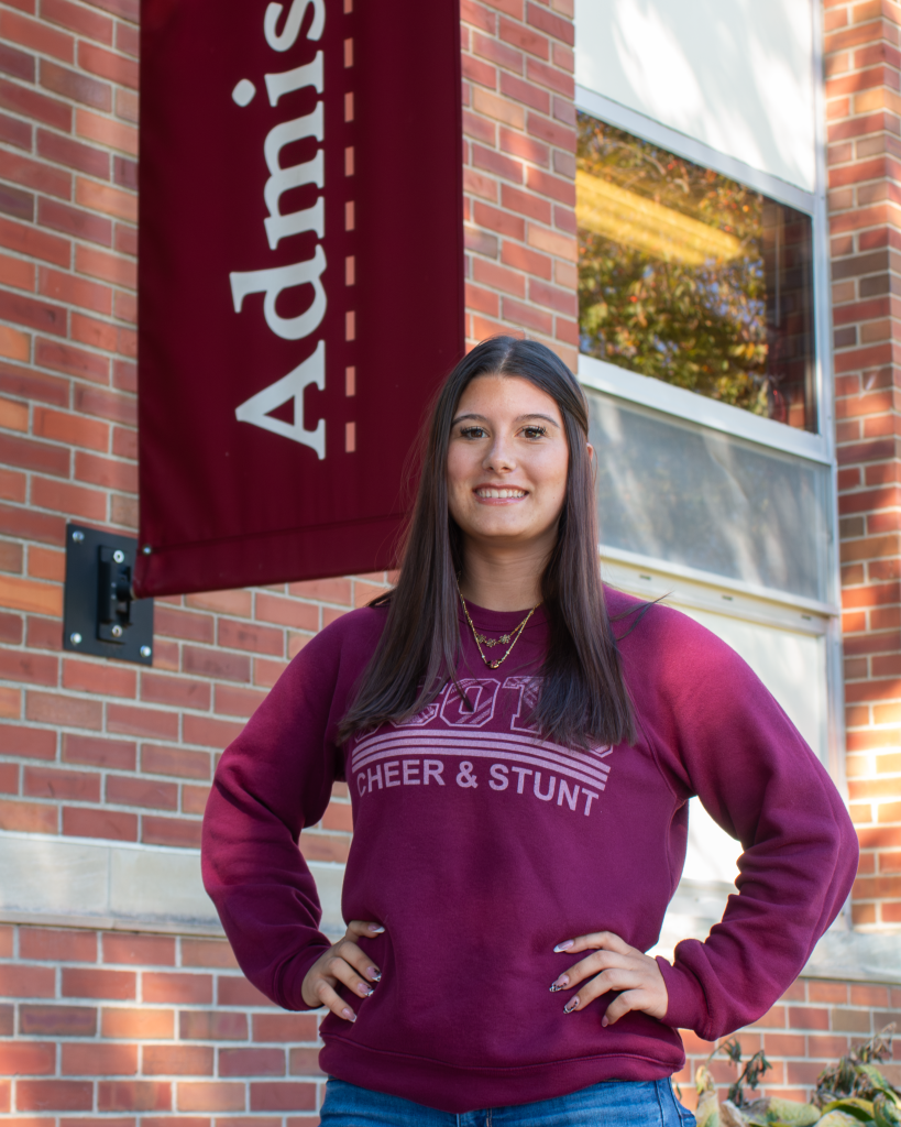 A young woman, one of our tour guides, wears a maroon cheer and stunt sweatshirt while standing in front of a brick building with an admissions sign.