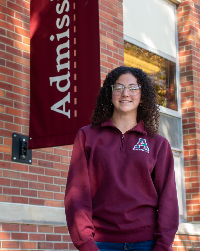 A student tour guide wearing glasses and a maroon quarter-zip pullover standing in front of an Admissions banner on a brick building.