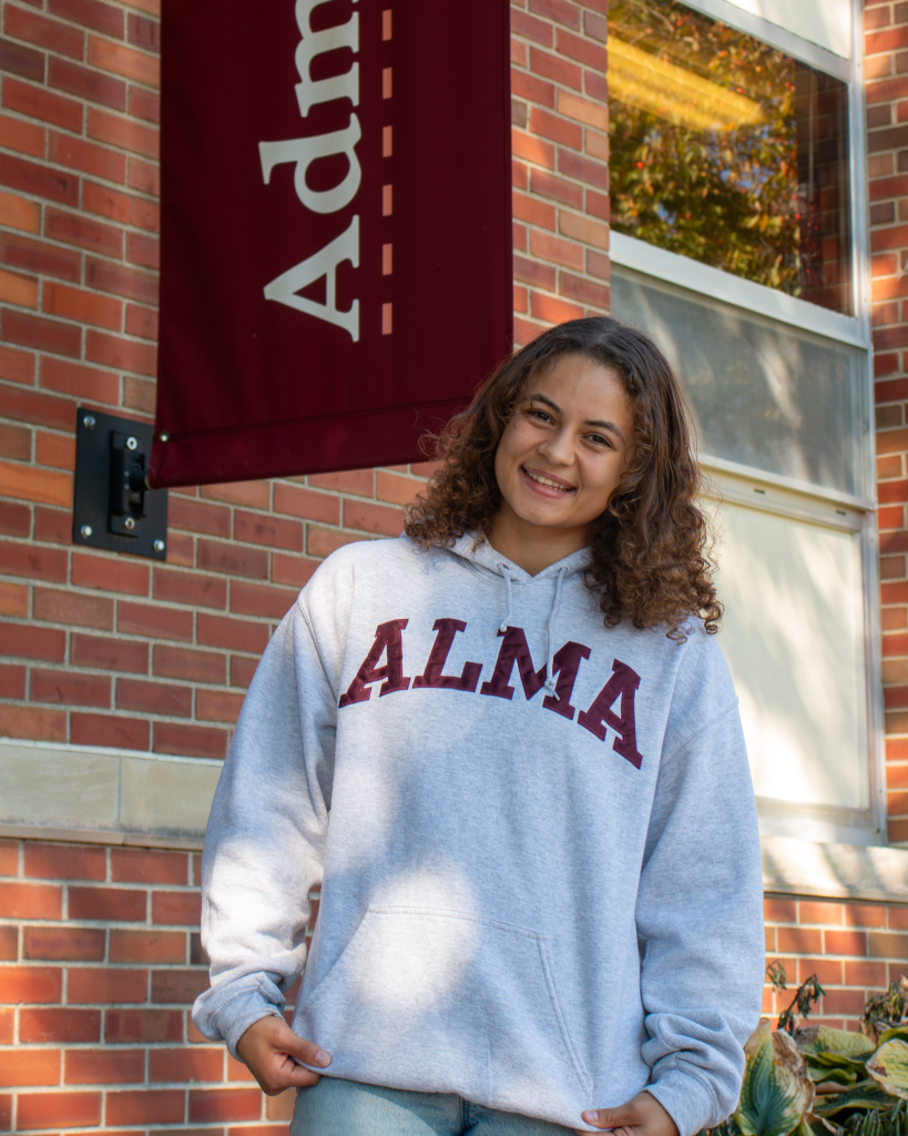 A smiling tour guide with curly hair wears a grey hooded sweatshirt with ALMA printed on the front, standing in front of a brick building with an Admissions sign.