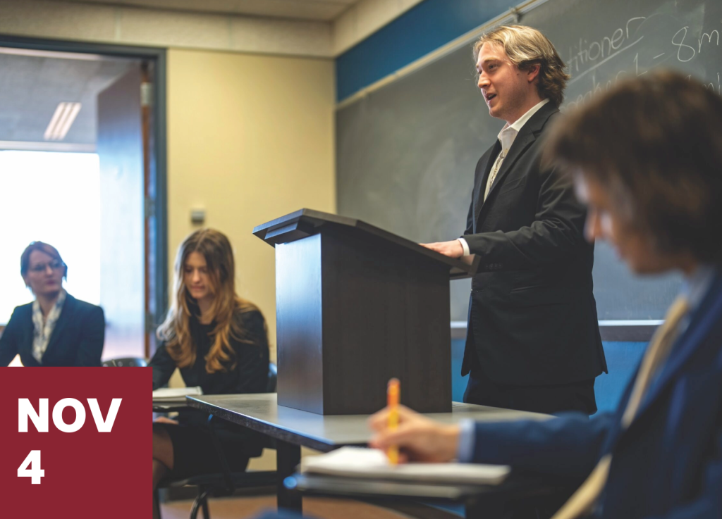 A man in a suit speaks at a podium in a classroom for a connect and visit session, with students seated at desks nearby and the text NOV 4 in a maroon box in the corner.
